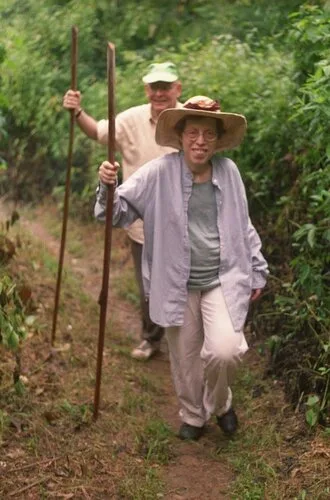 Ms. Bograd wearing a hat and hiking with her husband. They are holding walking sticks and there are trees all around them.